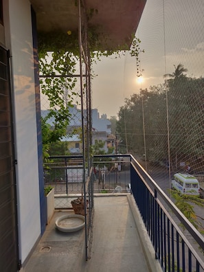 A balcony with a metal railing, a small planter, and a view of a street with a bus. - The Paperflower - A Wandertrails Stay (Puducherry)