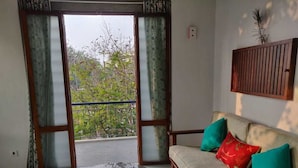 A living room with a sofa, a round table, and a view through a glass door to a balcony. - The Paperflower - A Wandertrails Stay (Puducherry)