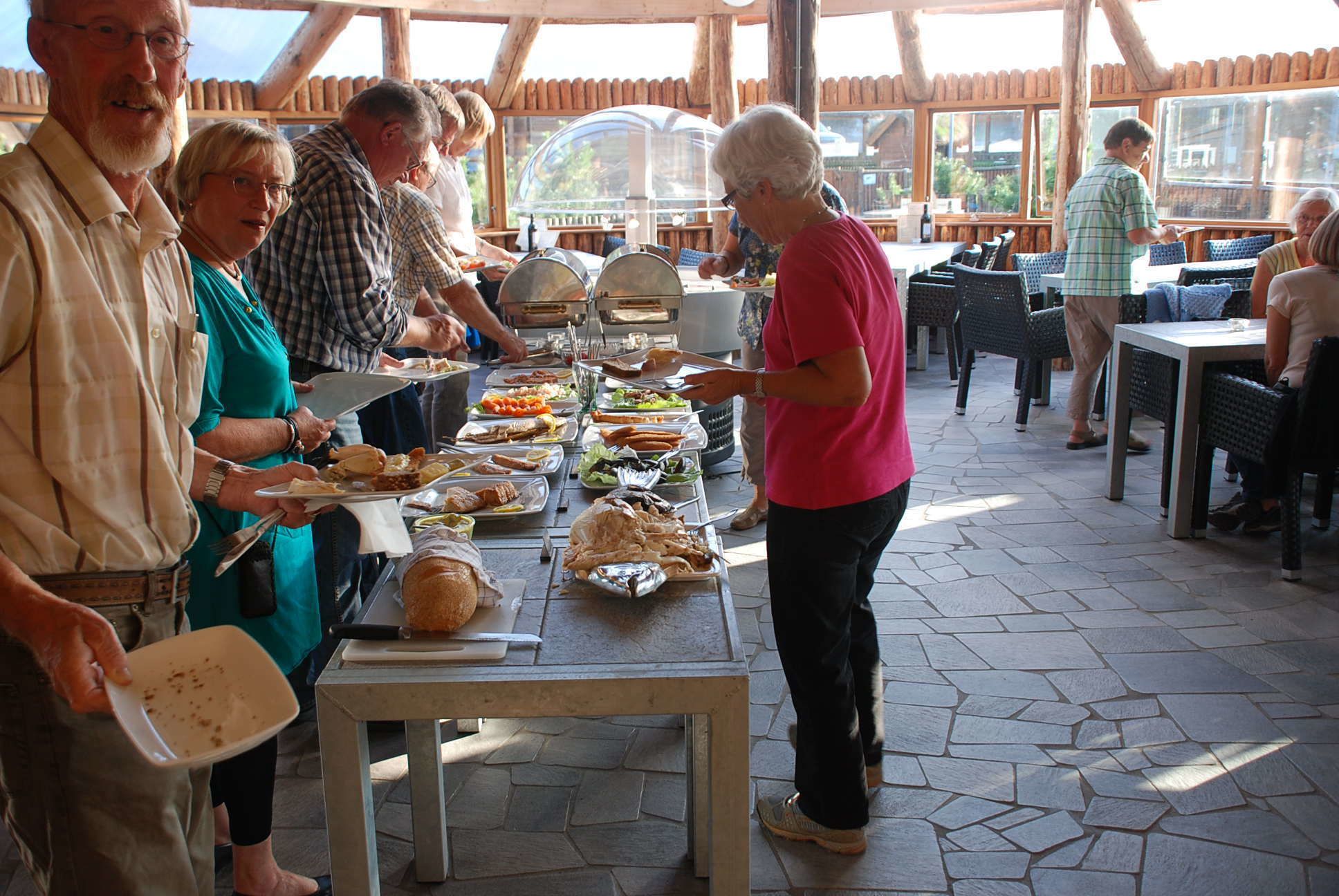 A buffet setup with various food items on trays, a table with a cutting board, and a group of people serving themselves.