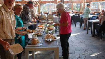 A buffet setup with various food items on trays, a table with a cutting board, and a group of people serving themselves.