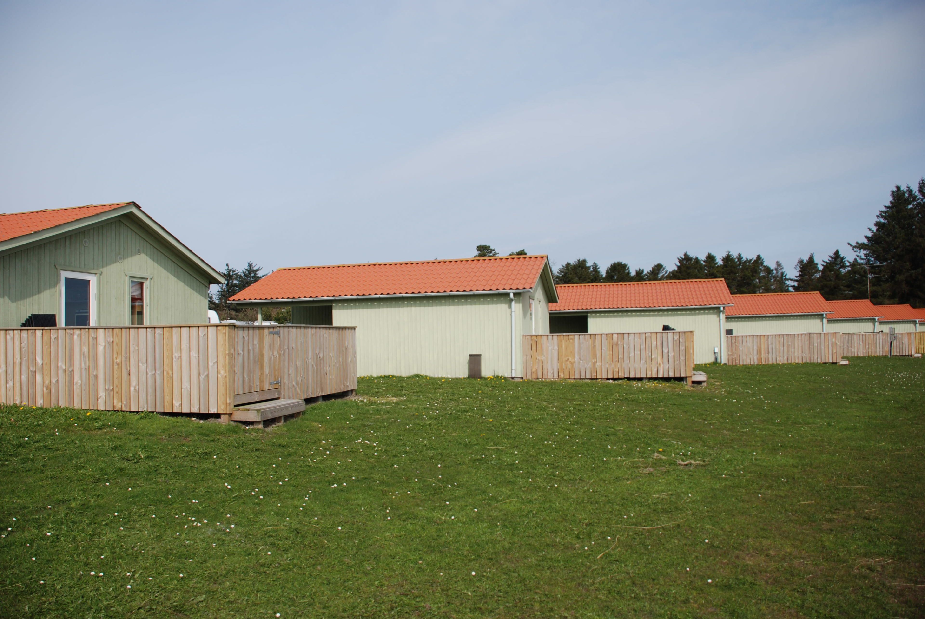Eine Reihe kleiner, einstöckiger Gebäude mit roten Dächern und Holzzäunen, eingebettet in eine grüne Wiese mit Bäumen im Hintergrund.