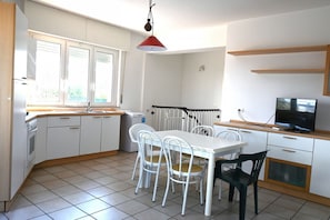 Une cuisine moderne avec des meubles blancs, une table à manger avec des chaises, un luminaire suspendu rouge et un téléviseur posé sur une étagère en bois.