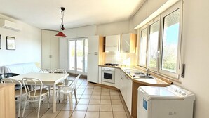 A modern kitchen with white cabinets, a dining table with chairs, a red pendant light, and a window with blinds.