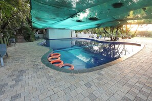 Una piscina con un borde de baldosas, rodeada de árboles y una zona con sombra.