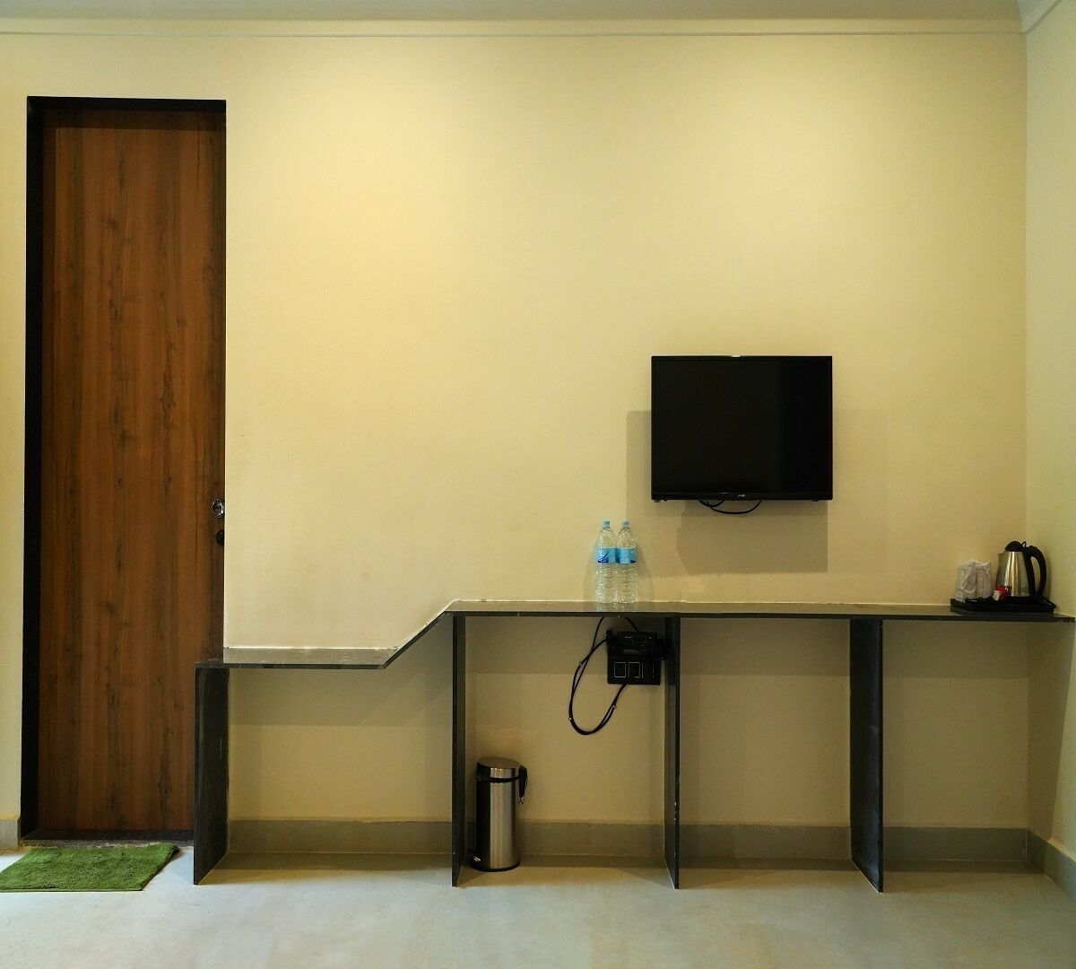 Una habitación con puerta de madera, un televisor de pantalla plana en la pared, una mesita con botellas de agua, un bote de basura y una cafetera.