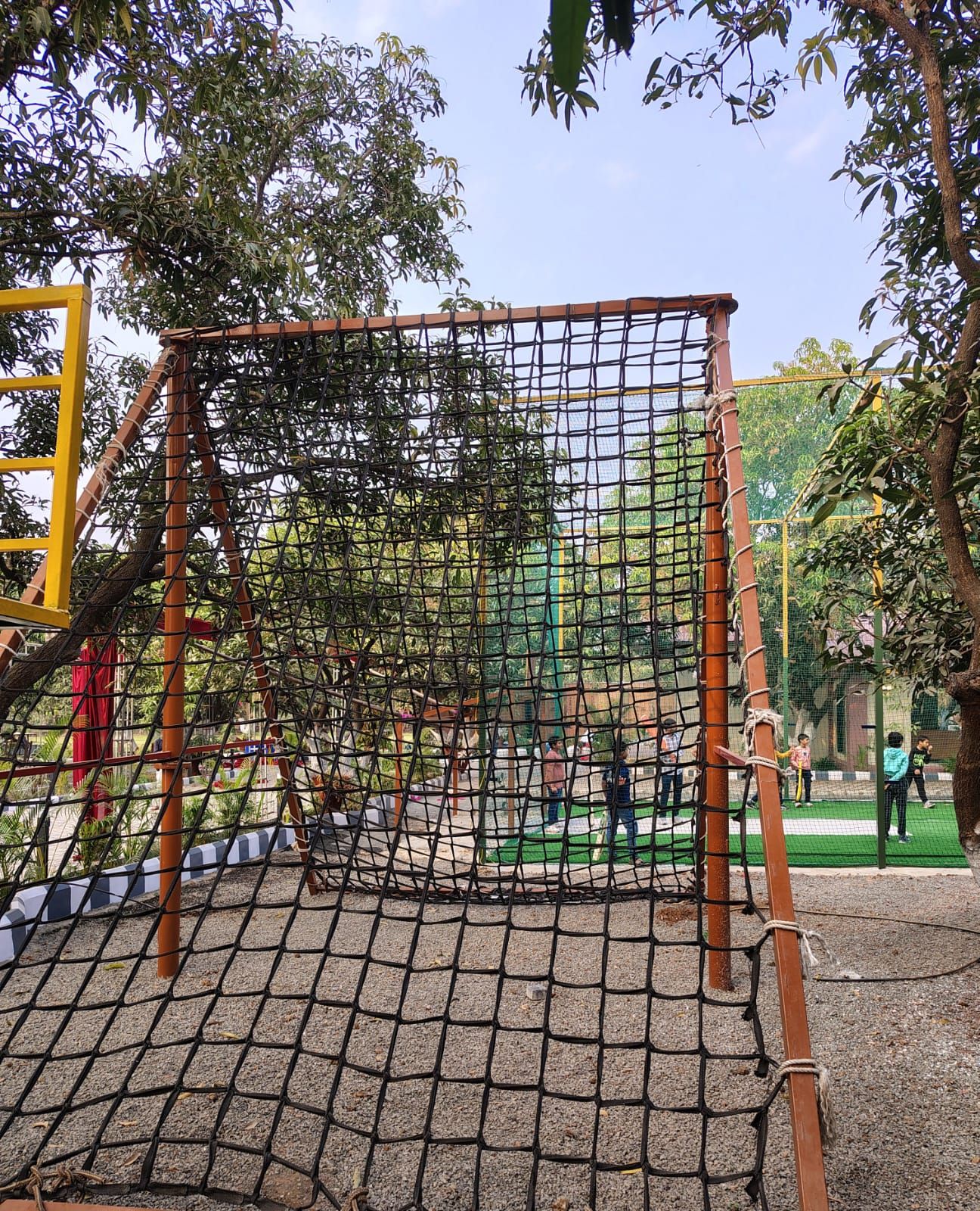 A playground with a netted climbing structure, surrounded by trees and a paved area.