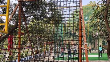 Un parque infantil con una estructura de escalada con red, rodeado de árboles y una zona pavimentada.