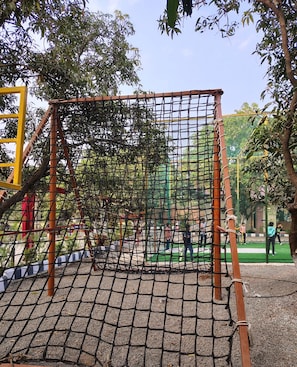 Un parque infantil con una estructura de escalada con red, rodeado de  árboles y una zona pavimentada.