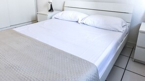Un llit amb llençols blancs, una tauleta de nit amb una làmpada i un armari blanc.