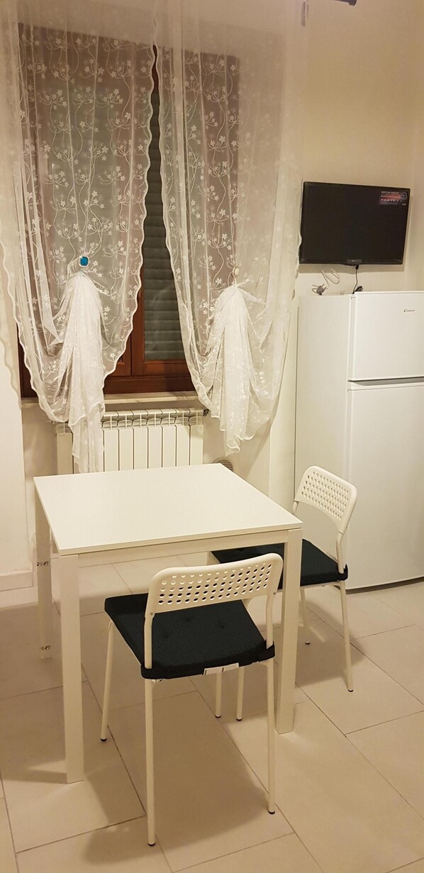 Una habitación pequeña y limpia con una mesa blanca y dos sillas, un refrigerador blanco y una ventana con cortinas traslúcidas.