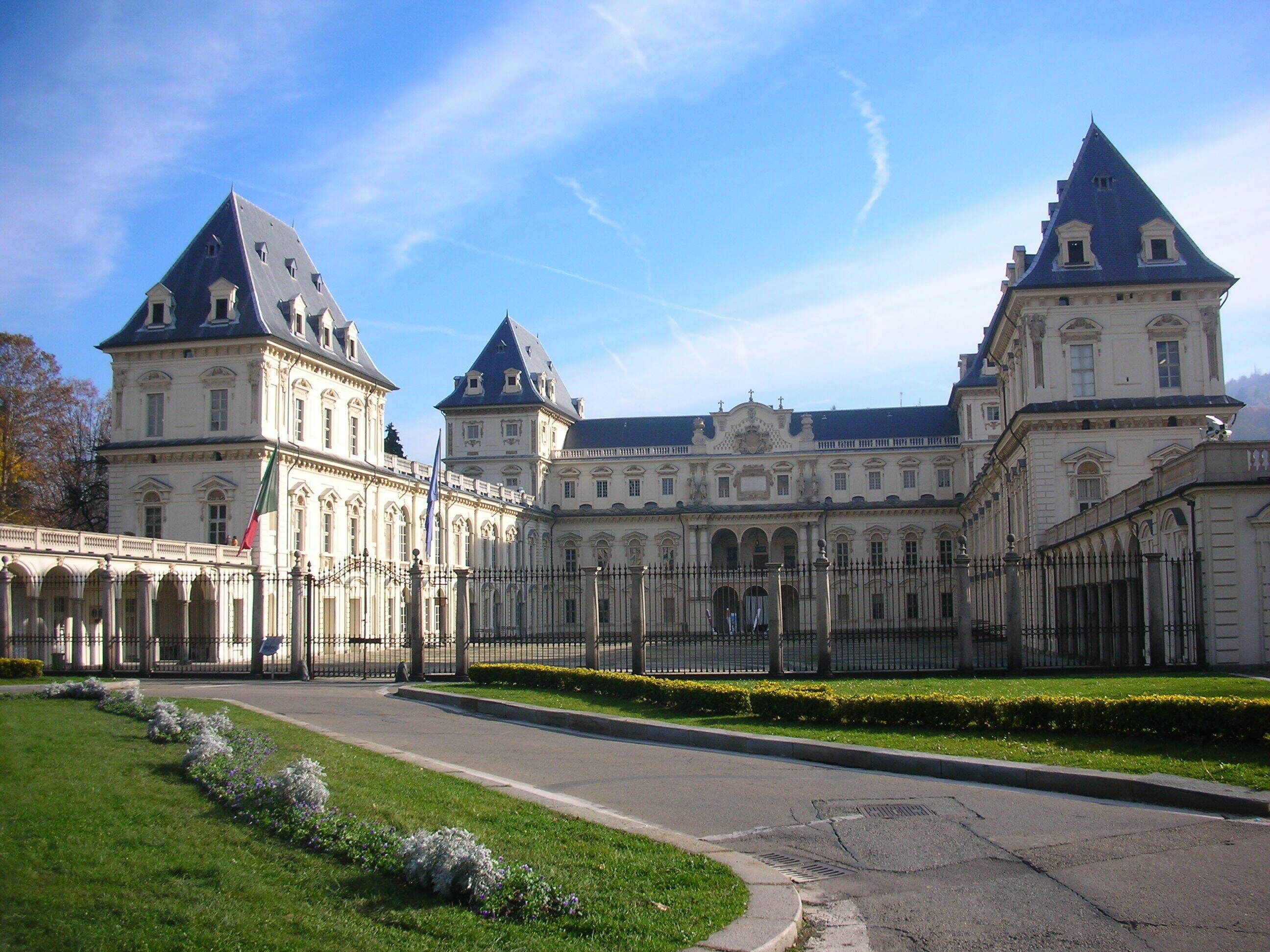 A large, historic building with multiple towers and a symmetrical facade, surrounded by a well-maintained lawn and a paved pathway.