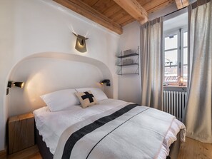 A bedroom with a large bed, wooden beams on the ceiling, a mounted animal head, and a window with curtains.