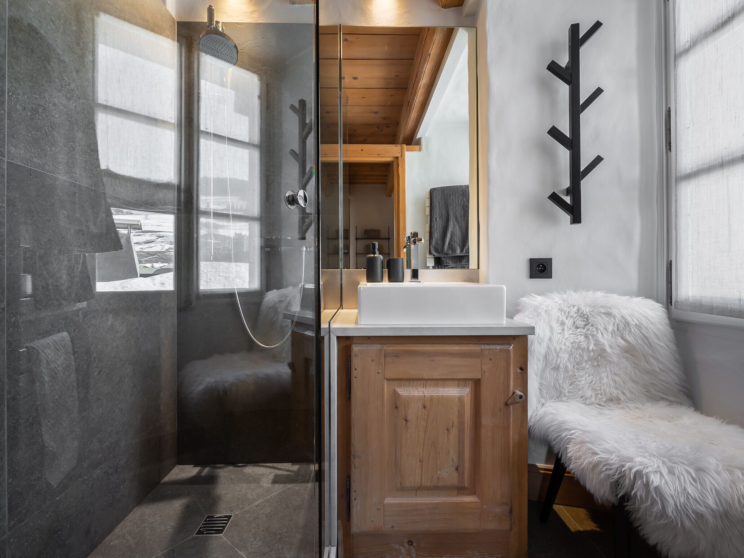 Een moderne badkamer met een douchecabine van glas, een houten kastje, een zachte witte bank en een grote spiegel.