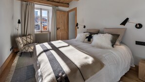 Un dormitorio con una cama grande, vigas de madera en el techo, una ventana con cortinas, una silla y dos lámparas de noche.