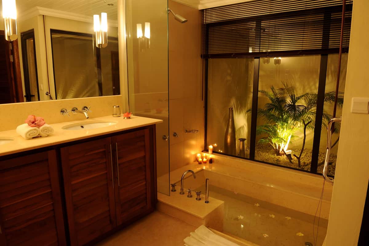 Un baño con un espejo grande, dos lavamanos, una ducha y una ventana con vista a palmeras.