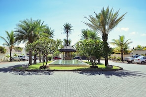 A paved courtyard with a central water feature, surrounded by palm trees and parked cars.