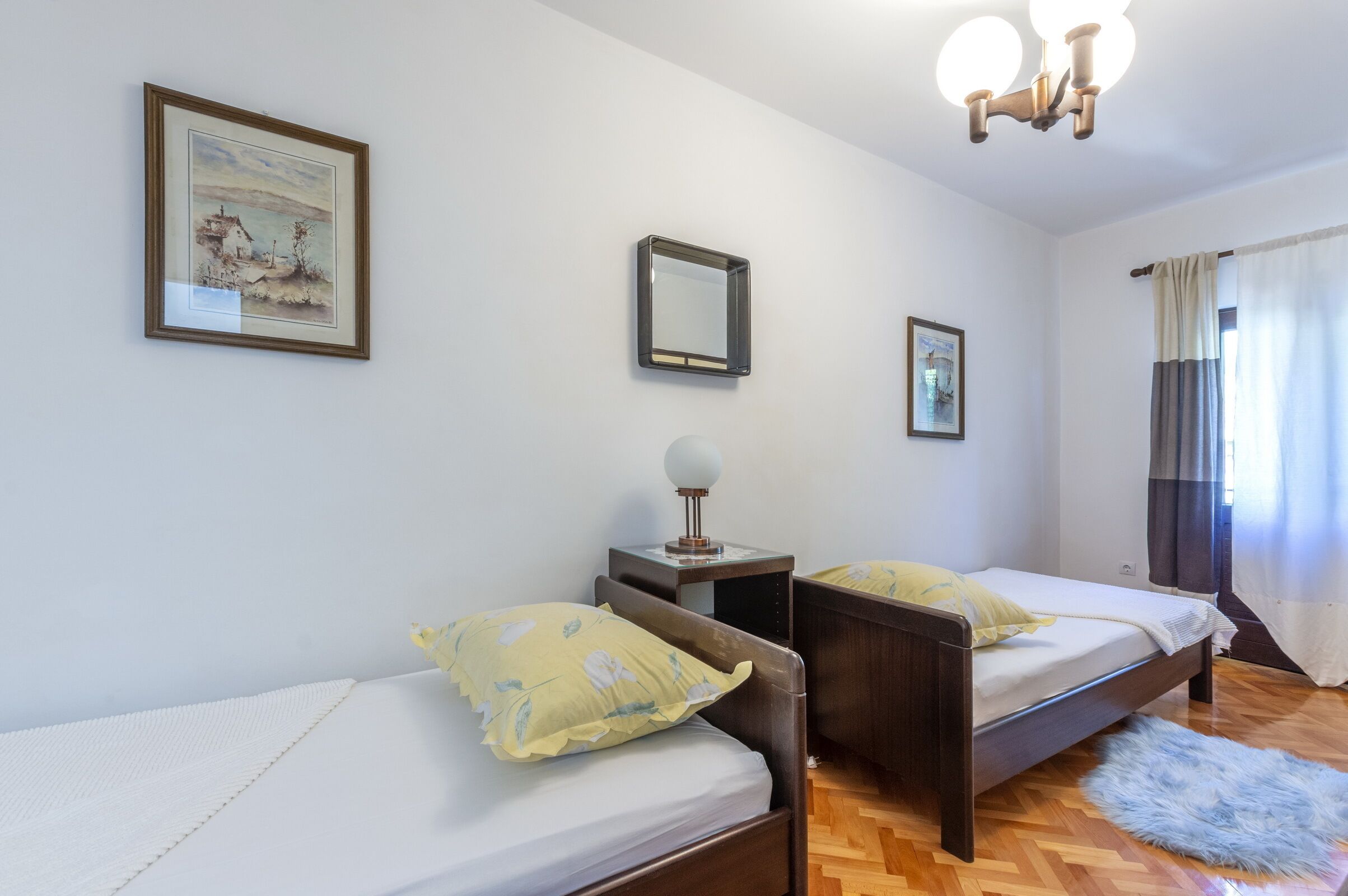 A bedroom with two single beds, a bedside table with a lamp, a framed picture on the wall, and a window with curtains.