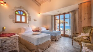 A bedroom with two beds, a wooden chair, a nightstand, a stone-framed window, and a view of the sea.