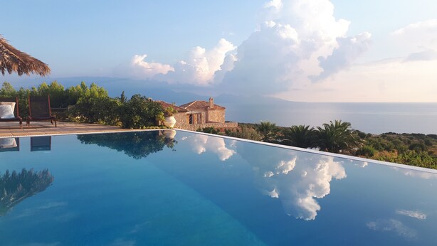 A clear blue infinity pool reflecting the sky and clouds, with a stone building and lush greenery in the background.