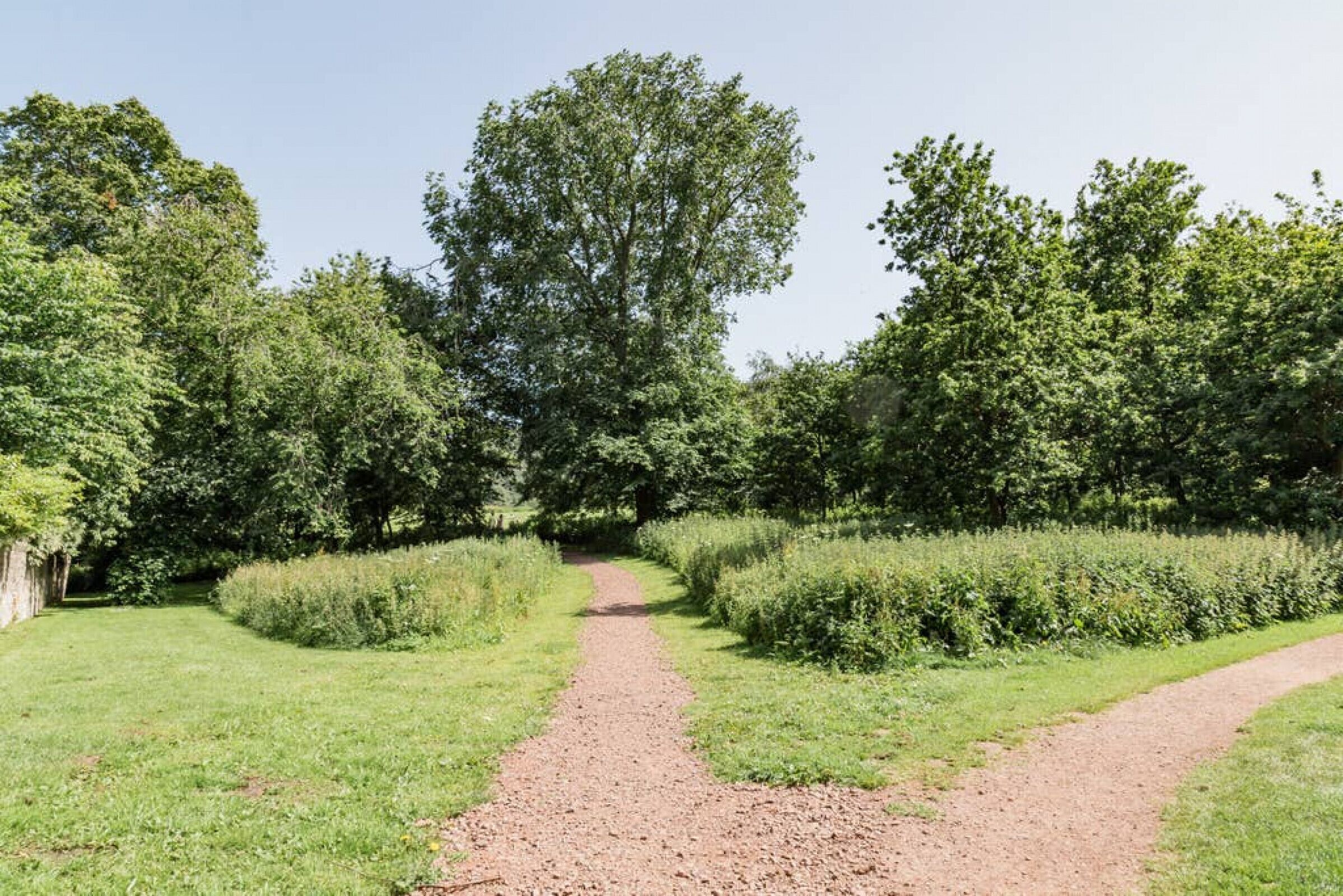 Un chemin de gravier traverse un parc où la végétation est luxuriante et où les arbres sont nombreux.