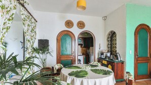 A dining area with a round table covered in a white tablecloth, surrounded by wooden chairs with floral upholstery. There are potted plants on the floor and a wooden cabinet with a green door in the background. - Homerez - Studio in Menton (Menton)