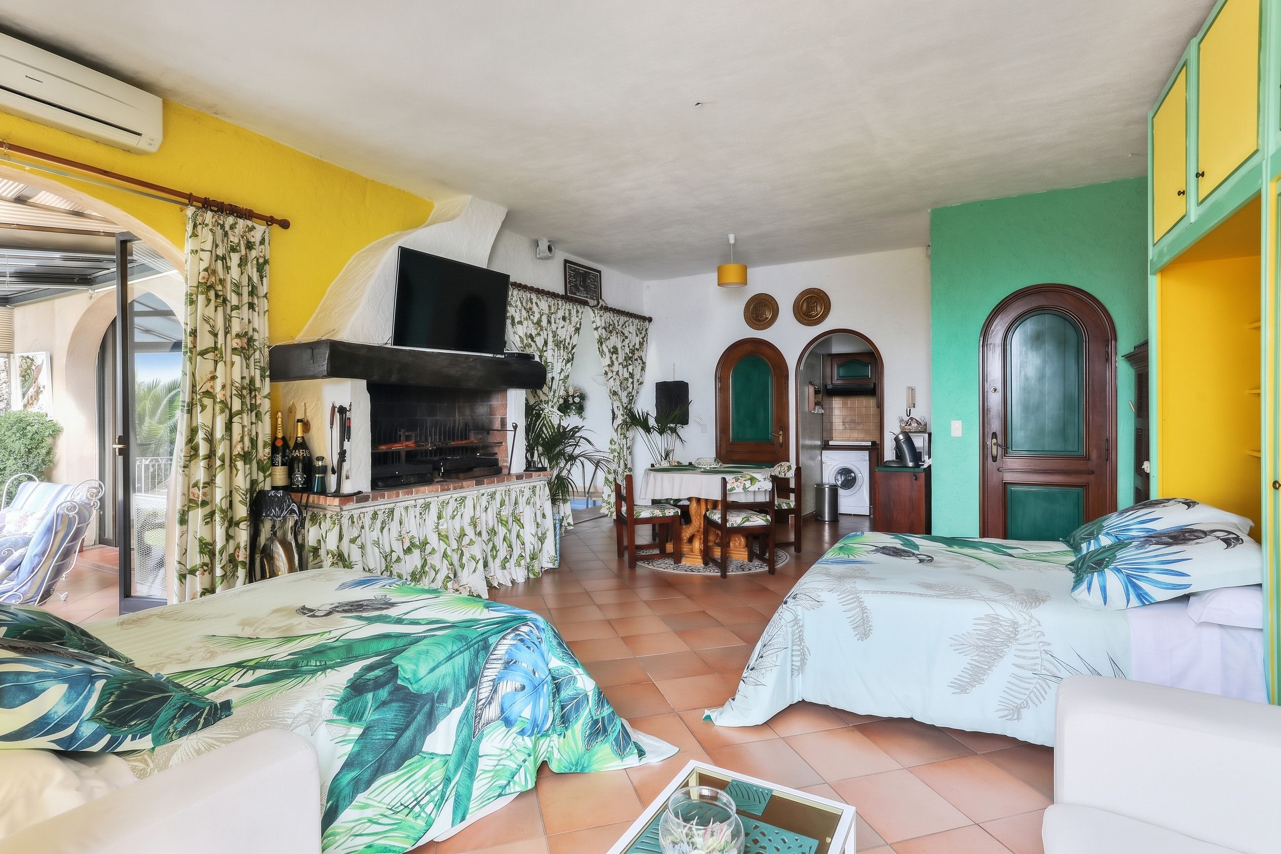 Une pièce avec une cheminée, un coin repas et un lit recouvert d’une couvre-lit à motifs tropicaux.