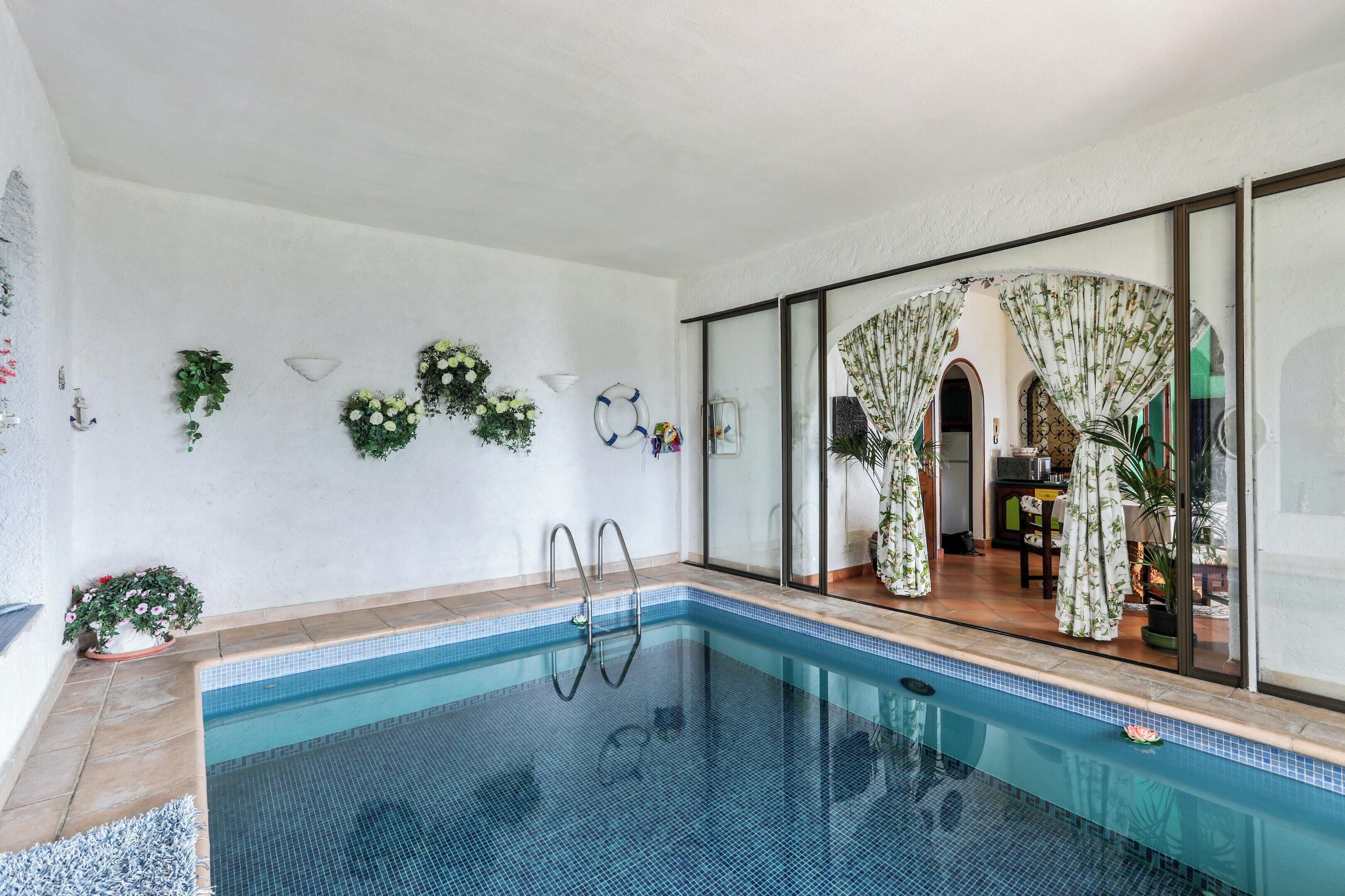 An indoor swimming pool with clear blue water, surrounded by a tiled floor and walls. There are potted plants and a glass door leading to a room with a patterned curtain.