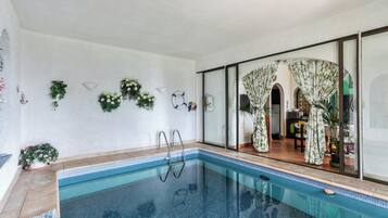 An indoor swimming pool with clear blue water, surrounded by a tiled floor and walls. There are potted plants and a glass door leading to a room with a patterned curtain.