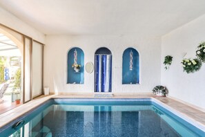 Una piscina con piso y paredes de cerámica, una ventana con vista a follaje, y una puerta con cortina azul.