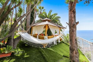 A secluded outdoor hammock bed with a thatched roof, surrounded by lush greenery and palm trees, overlooking a clear view of the ocean. - Homerez - Studio in Menton (Menton)