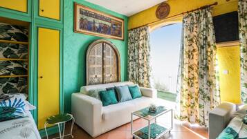 Una stanza con un divano bianco, un tavolino e una poltrona verde. È presente un'ampia finestra con tende e vista sull'esterno.
