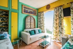A room with a white sofa, a small table, and a green chair. There is a large window with curtains and a view of the outdoors. - Homerez - Studio in Menton (Menton)