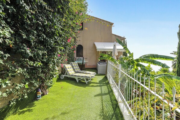 Un patio con césped artificial, dos tumbonas, un parasol blanco y una valla blanca adornada con plantas.