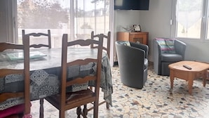 A living room with a dining table, chairs, a sofa, a TV, and a window. - Single-Story House: Central Location, 150m from Beach, Terrace, Parking (Jullouville)