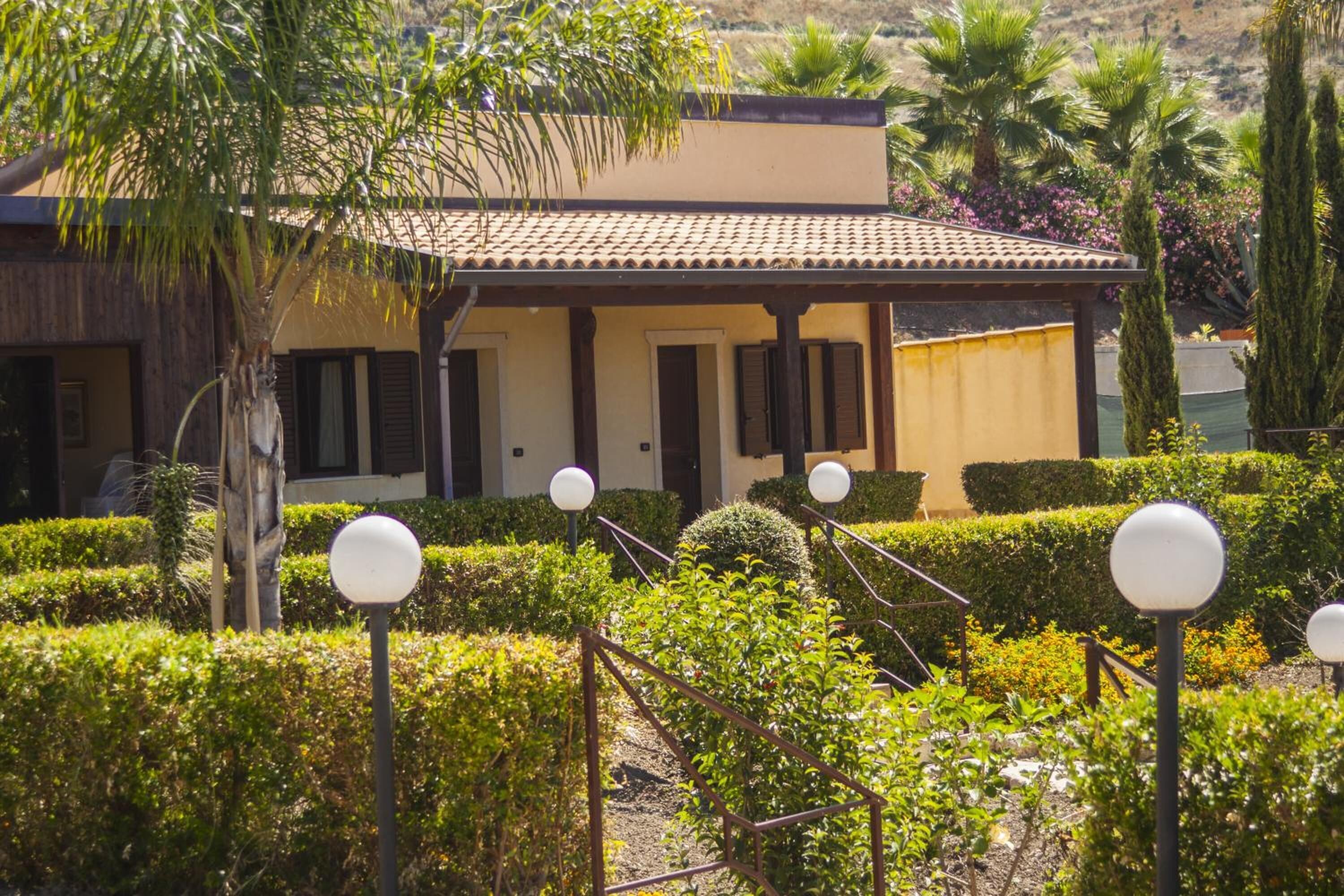 A building with a tiled roof, a door, and windows with shutters. There are hedges and a pathway with spherical lights.