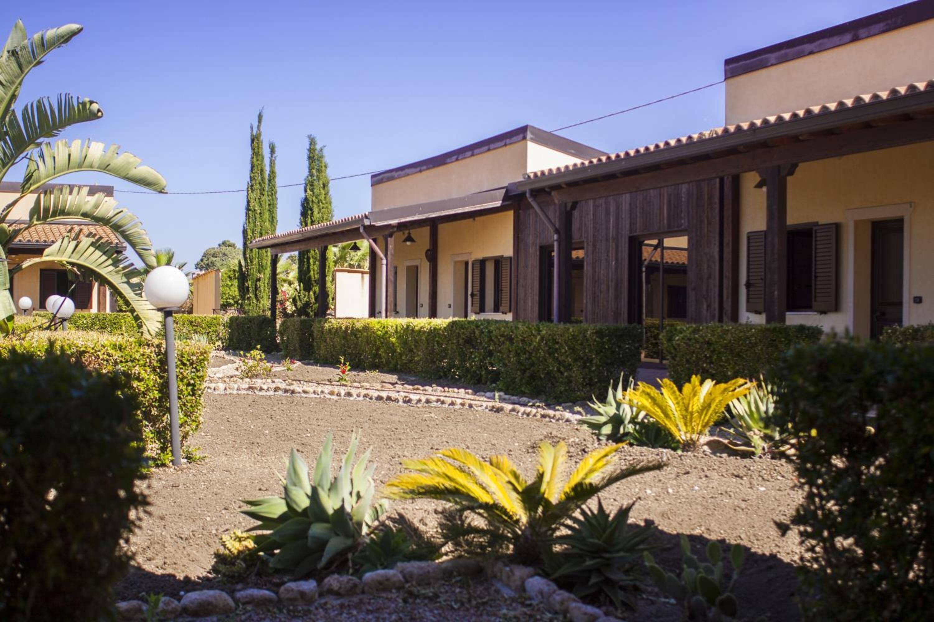 A building with a covered patio area, a garden with various plants, and a clear sky.