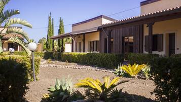 A building with a covered patio area, a garden with various plants, and a clear sky.