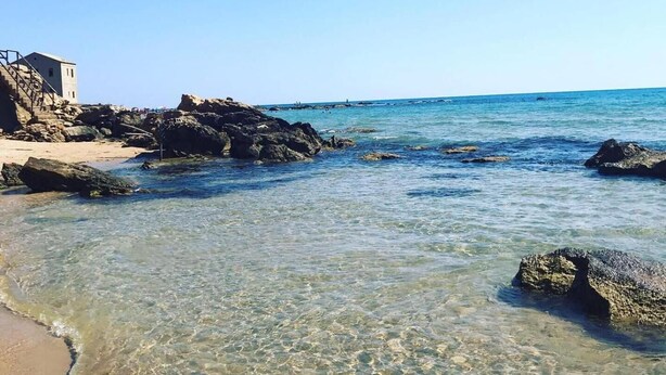 Una playa con aguas claras y poco profundas, afloramientos rocosos y un edificio a la izquierda.