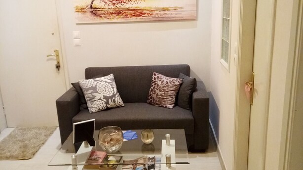 Ein Wohnzimmer mit grauem Sofa, einem Glastisch mit Zeitschriften, einem Gemälde an der Wand und einer Tür rechts.