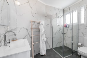 Un baño moderno con lavamanos blanco, paredes de mármol, cabina de ducha de vidrio y un toallero de madera.