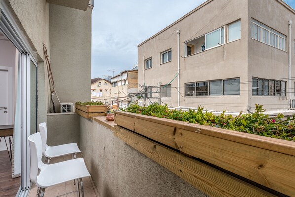 Un balcón con una jardinera de madera, sillas blancas y vistas a edificios cercanos.