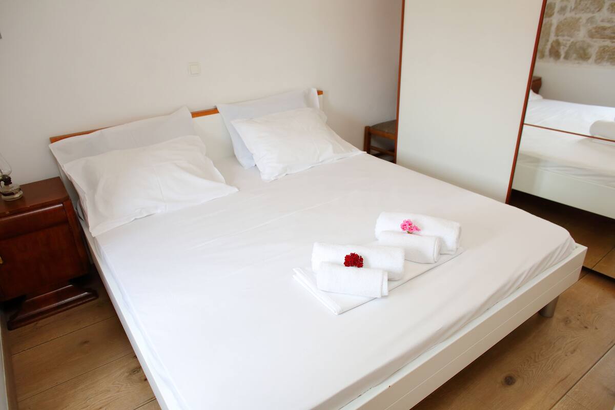 A neatly made bed with white linens and towels, a wooden nightstand with a lamp, and a mirror reflecting the bed.