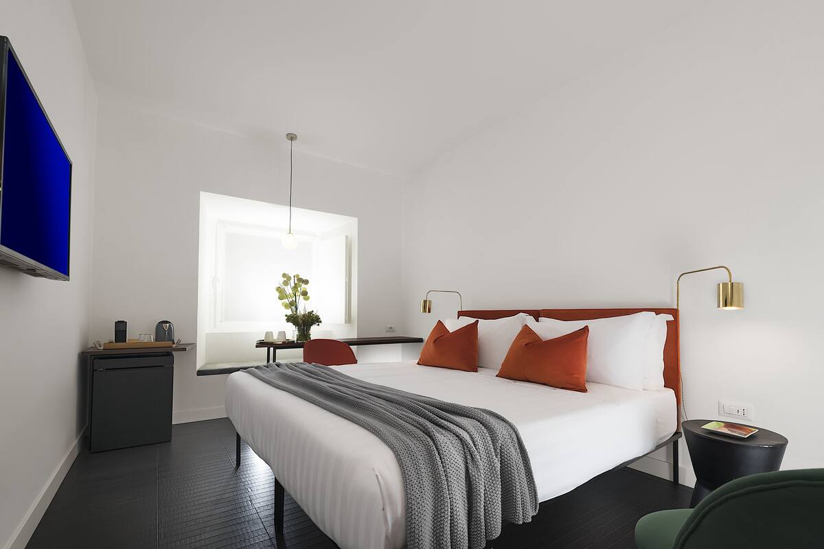 A modern hotel room with a large bed, a desk, a chair, a small table, and a TV on the wall.
