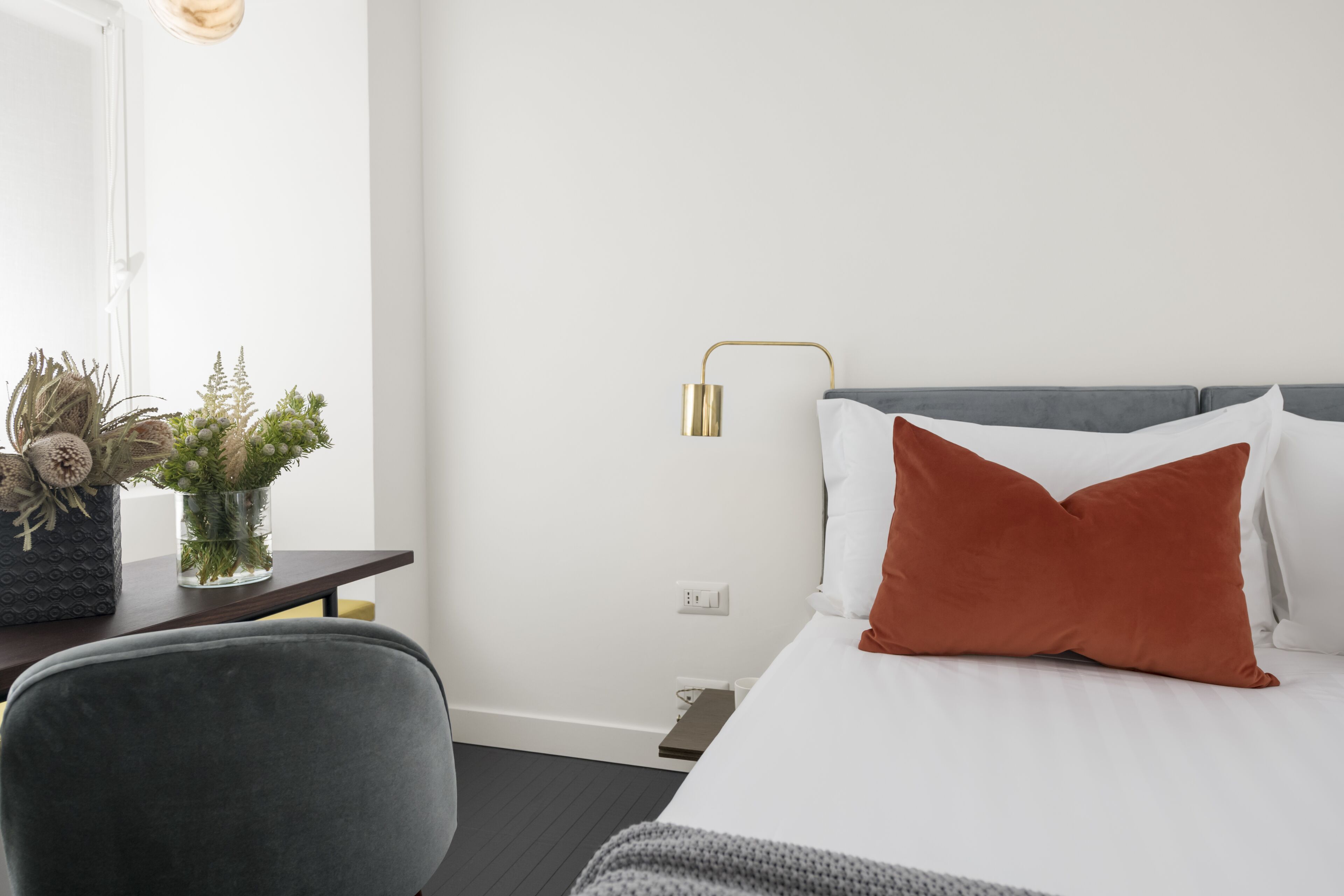 A hotel room with a bed, a desk with a vase of plants, a chair, a bedside table, and a wall-mounted lamp.