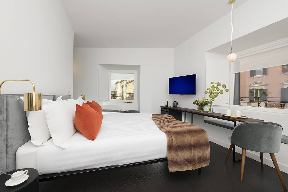 A modern hotel room with a bed, a desk, a TV, and a window with a city view.