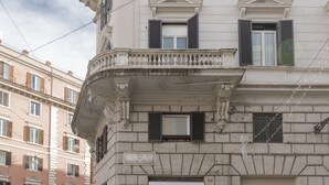 A corner building with classical architecture, including a balcony with balustrades, multiple windows with shutters, and a sign indicating a street name. - Maison Ottavia (Rome)