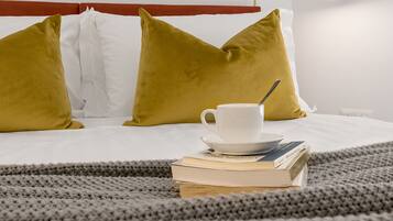 Una cama impecablemente arreglada con almohadas blancas y mostaza, una taza de café y una pila de libros sobre una manta tejida gris.