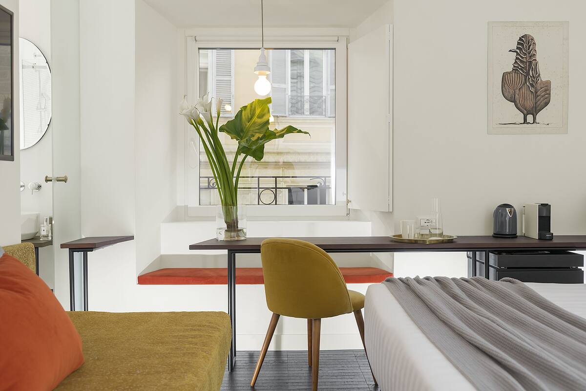 A modern hotel room with a bed, a desk with a chair, a window with a view, and a plant.