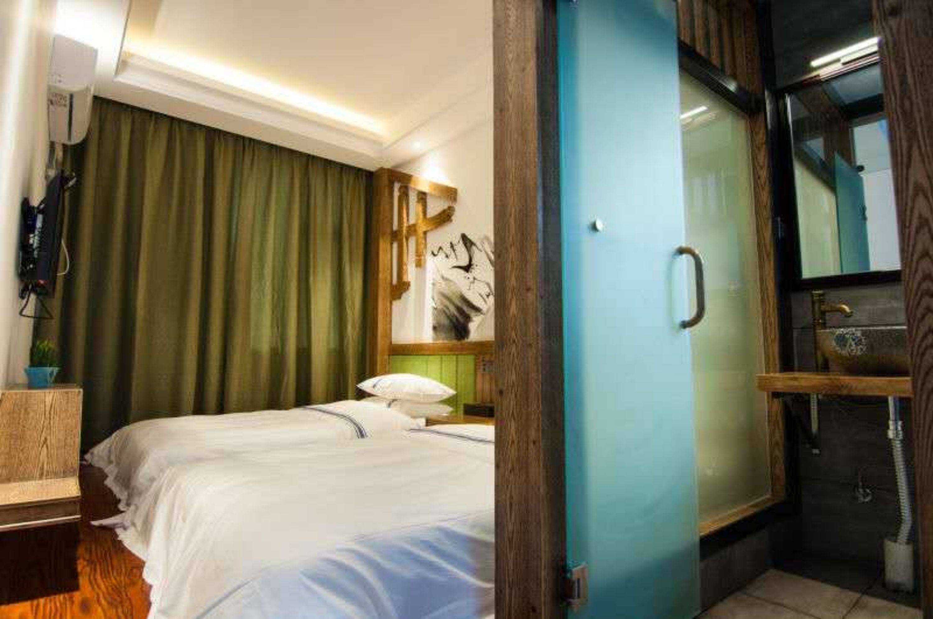 A hotel room with a bed, a green curtain, a wooden headboard, a painting, a bathroom with a sink and mirror.