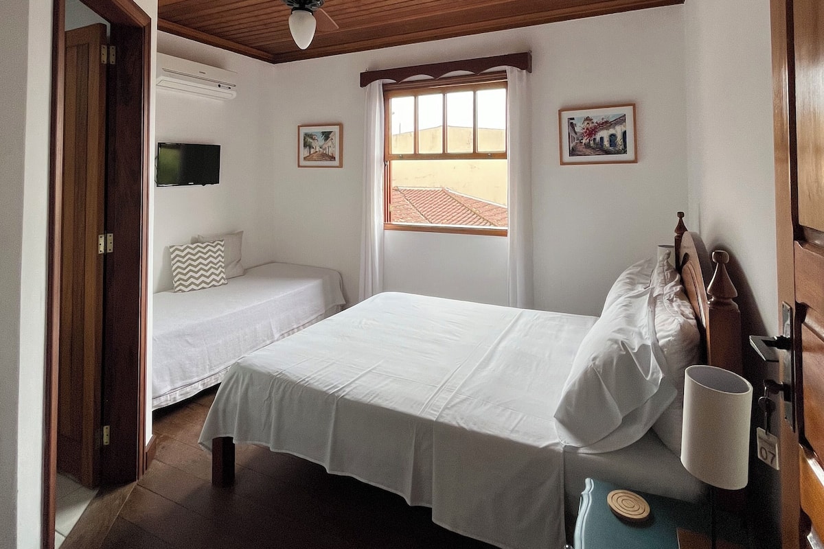 A bedroom with two beds, a wooden ceiling, a window with a view, and a bedside table with a lamp.